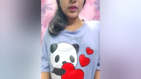 Video of a young woman with medium brown skin, wearing a light blue t-shirt featuring a panda holding a red heart, surrounded by red hearts, against a blurred pink and purple background.