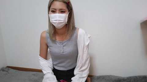 Video of a young woman with light skin, straight, shoulder-length blonde hair, wearing a white face mask, gray tank top, white shirt draped off shoulders, and black skirt, sitting on a gray couch against a plain white wall.