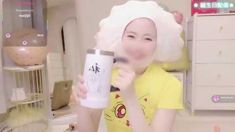 Media: Video of a woman with a large white shower cap, yellow t-shirt, holding a white water bottle, standing in a cluttered, brightly lit room with a water dispenser and white furniture.