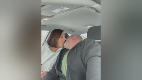 Video of an intimate moment in a car; a woman with short brown hair and a white shirt leans in to kiss a bald man in a gray jacket.