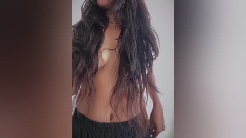 Video of a topless, dark-skinned woman with long, wavy black hair, wearing black shorts. Background is a gradient of brown and beige.