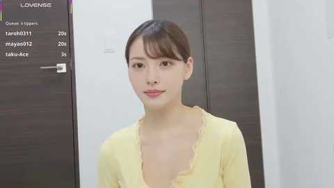 Media: Video of a young Asian woman with light skin, brown hair in a simple updo, wearing a yellow blouse with ruffled collar, standing in front of a dark wood cabinet with a white background.