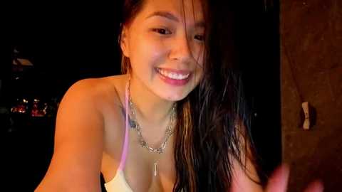 A video of a young Asian woman with wet, dark hair, smiling widely, wearing a white spaghetti-strap top, standing indoors at night.