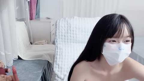 A video of an Asian woman with long black hair and a surgical mask, sitting on a white textured chair in a sparsely furnished room with light walls and a blue carpet.