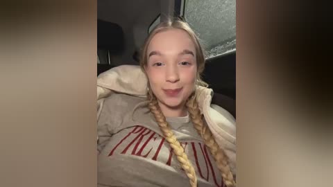 Video of a smiling young woman with blonde braids, wearing a grey hoodie with \"Princess\" in red text, sitting in a car with a foggy window background.