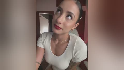 Media: Video of a light-skinned woman with medium-length dark hair, wearing a white top, red lipstick, and a necklace. She gazes upward, tattoos visible on her arm, in a dimly lit room.