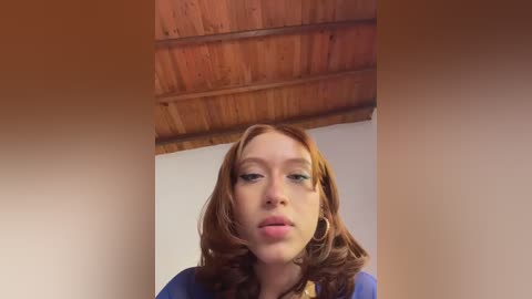 Video of a fair-skinned redhead with shoulder-length wavy hair, wearing hoop earrings, and a blue top, set against a wooden ceiling.