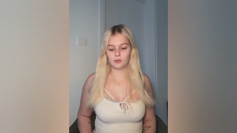 Media: A video of a young Caucasian woman with long, straight blonde hair, wearing a beige tank top with thin straps, standing in a plain, dimly lit room with neutral-colored walls.