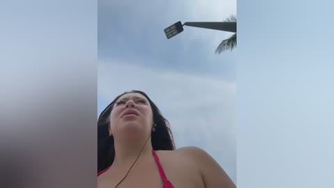 Video of a young woman with fair skin, dark hair, and a red bikini, looking up at a streetlight. Cloudy sky in the background, with a palm tree visible.