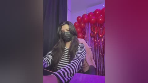 Video of a woman with long dark hair, wearing a black mask, striped sweater, and jeans, sitting on a bed with red balloons and black curtains in the background.