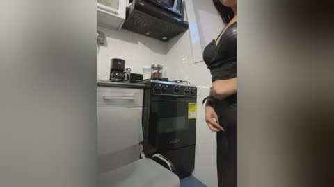 Video of a small, modern galley kitchen with a woman in a black dress standing near a black stove and microwave. The space is clean, with white walls and a gray floor.