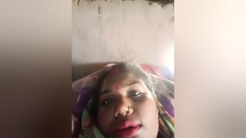 Media: A close-up video of a young woman with a light brown complexion and green eyes, wearing a colorful, patterned headscarf, with Holi-colored powder smeared on her face. The background is a plain, light beige wall.