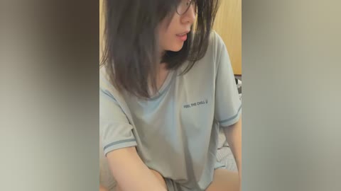 Media: Video of a young East Asian woman with shoulder-length black hair, wearing a gray t-shirt and black glasses, sitting indoors, partially obscured by a blurred background.