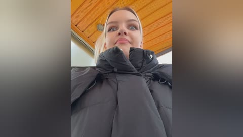 Video of a blonde woman with light skin, wearing a black puffy jacket, standing indoors under a wooden ceiling with orange panels. She has a neutral expression and is slightly out of focus.