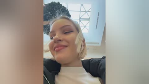 Video of a young woman with blonde hair, wearing a black jacket over a white shirt, smiling, in a brightly lit room with a window and a decorative black fan.