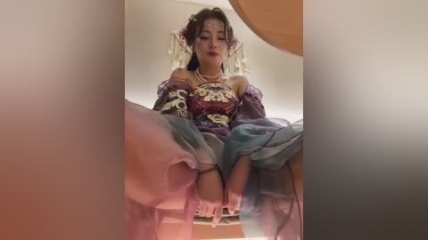 A video of a woman in an elaborate, ornate outfit with gold accents, sitting provocatively with legs spread, surrounded by blurred, pastel-colored backgrounds.
