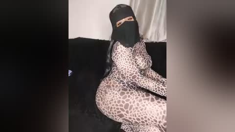 Video of a woman with long black hair, wearing a tight leopard-print bodysuit, black mask, and sitting on a black couch against a white wall, creating a dramatic, mysterious atmosphere.