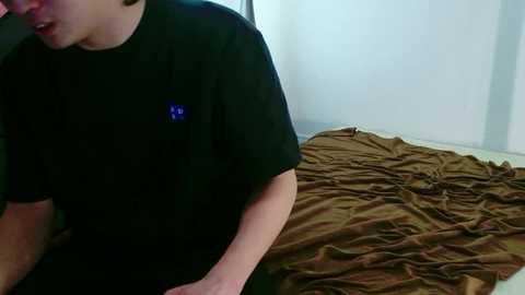 Video of a young man in a black T-shirt with a blue logo, sitting on a bed with a rumpled brown blanket. The background features a plain white wall.