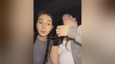 A video features two women with long hair in braids, one with fair skin and a gray top, the other with a tan complexion and a gray jacket. They stand side by side, both looking slightly off-camera, creating a blurred, intimate, indoor setting.