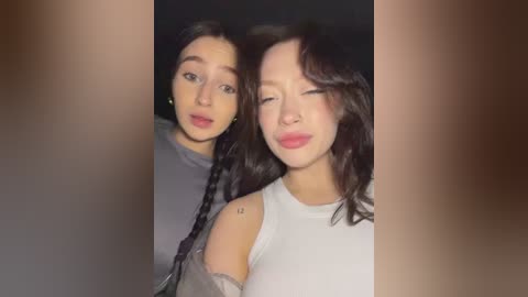 Video of two women with long dark hair, one in a gray top, the other in a white top, both smiling, against a blurred, dark background.