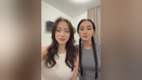Video of two young women with fair skin, long brown hair, and light makeup, wearing casual white and gray tops, standing indoors with beige curtains in the background.