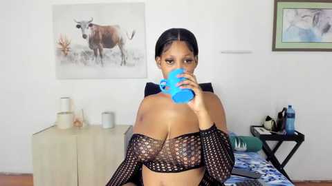 Media: A video of a Black woman with dark skin, long hair, and large breasts, wearing a black fishnet top, drinking from a blue water bottle in a modern, minimalistic bedroom.