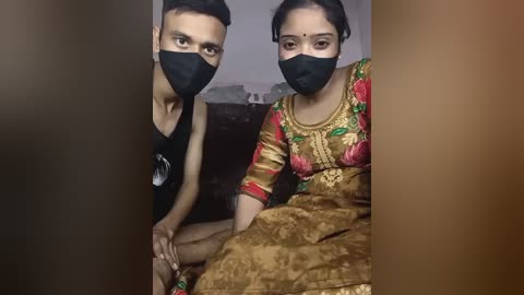 Media: Video of a young South Asian couple, both wearing black face masks and traditional clothing; he in a black tank top, she in a floral-patterned gold sari.