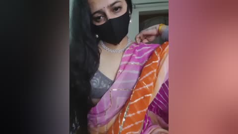 Media: Video of a South Asian woman with medium skin tone, long black hair, wearing a black mask, grey bra, and a colorful saree, indoors, blurred background.