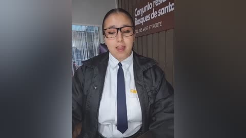 Video of a Latina woman in a dark jacket over a white shirt, blue tie, and glasses, standing in a dimly lit office with Spanish text on a sign and a partially visible co-worker.