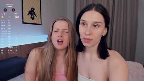 Video of two young women with light skin, one with long blonde hair, the other with straight black hair. They wear strapless tops, appear to be in a bedroom setting with a TV displaying a video game.