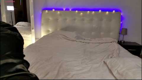 Video of a modern bedroom with a tufted white headboard, a bedside table, and a lamp, lit by blue LED strip lighting. The bed is unmade, with white sheets and a dark jacket partially visible.