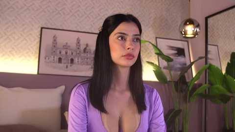 Video of a young woman with straight black hair, wearing a low-cut purple top, sitting indoors with a beige couch, potted plants, and framed black-and-white art on the wall.