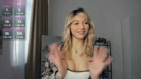 Video of a smiling, light-skinned woman with wavy blonde hair, wearing a white tank top, in a home with a digital workout display, a mirror, and a shelf with plants.