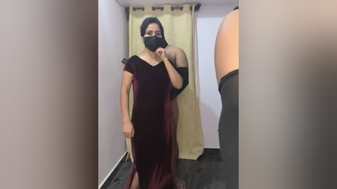 Video of a woman in a maroon velvet dress with a black face mask, standing in a fitting room with beige curtains and a gray floor.