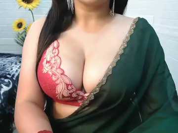 Video of a woman with medium skin tone, wearing a red lace bra with floral embroidery, green sari, and large silver earrings. Background includes white brick wall and sunflowers.