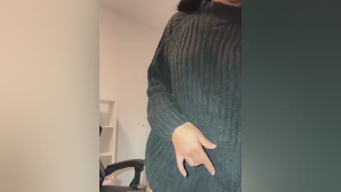 Video of a person in a dark green, ribbed-knit sweater, standing in an office with a white chair and a beige wall in the background. The person is partially obscured, with only their torso visible.