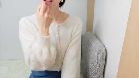 Media: Video of an Asian woman with light skin, wearing a white, fluffy cardigan and blue skirt, touching her lips, seated on a grey chair against a white wall.