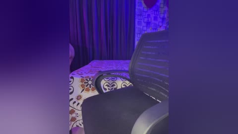 Media: Video of a dimly lit bedroom with a bed covered in a floral-patterned bedspread, featuring orange and purple hues. A black chair is positioned in the foreground, partially obscured by a blue-purple light effect.