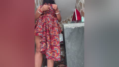 Video of a woman with medium-dark skin, wearing a colorful patterned dress, standing in a rustic room with stone walls and a gray refrigerator. She's holding the dress up, revealing her legs, and her wrists are bound together with red and white cords.