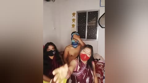 Video of two Asian women in a dimly lit room with a man wearing a blue mask, black face covering, and red shirt. Background shows white walls, a window with blinds, and a patterned rug.