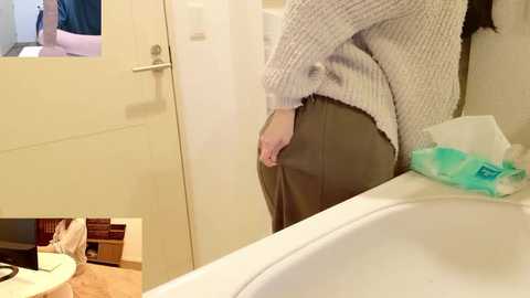 Video of a woman in a beige knit sweater and grey skirt, standing in a bathroom with a white sink and countertop, looking in a mirror. Background includes a closed door and wooden floor.