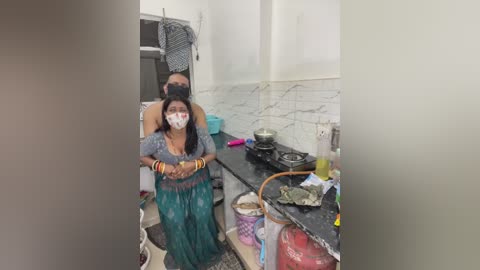 Video of a woman with a cloth over her head, wearing a mask, sitting on a kitchen counter amidst cooking utensils and a gas stove, surrounded by white tiled walls.