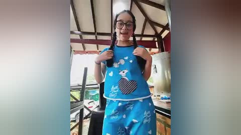 Video of a smiling Asian woman with glasses and braided hair, wearing a blue apron with a cartoon pig design, standing in a rustic outdoor kitchen with wooden beams and a water dispenser.