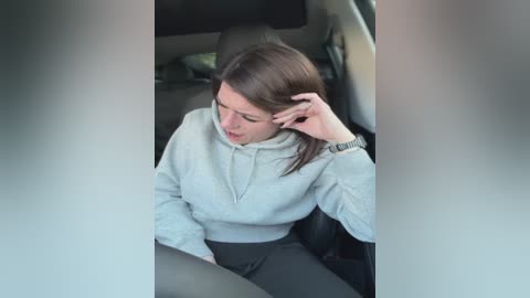 Video of a woman with fair skin, straight brown hair, and wearing a grey hoodie and dark pants, sitting in a car seat, looking stressed, touching her forehead.