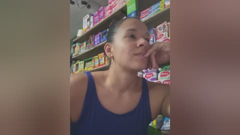 Video of a young woman with light skin and dark hair in a ponytail, wearing a blue tank top, standing in a brightly lit convenience store with colorful candy and snack displays in the background.