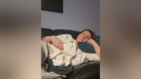 Video of a fair-skinned woman with brown hair, lying on a dark gray couch, wearing a white robe and gray pajama pants, eyes closed, looking relaxed.