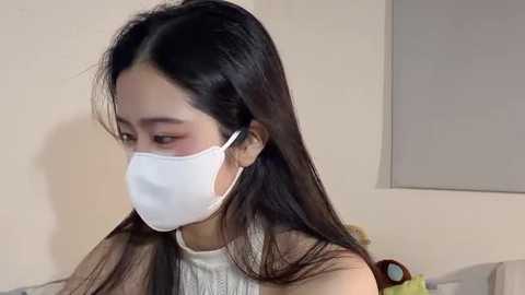 Video of an East Asian woman with long black hair and fair skin, wearing a white mask and sleeveless white top, indoors with beige walls and a gray window shade.