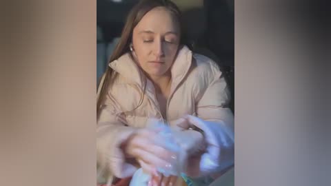 Video of a young woman with long brown hair, wearing a white puffer jacket, eyes closed, holding a lit candle in a dimly lit room.