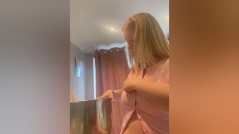 A video captures a blonde woman in a pink shirt stirring a metallic pot in a dimly lit kitchen with beige walls and red curtains.