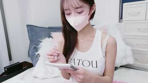 Media: Video of an East Asian woman with long, straight brown hair, wearing a white tank top with \"UOU\" in black, a white mask, and a white feather boa. She's writing in a notebook, in a minimalist bedroom with a bed and white dresser.